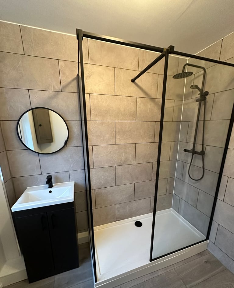 Modern bathroom with black-framed glass shower enclosure, white basin vanity, circular mirror, and beige tile walls